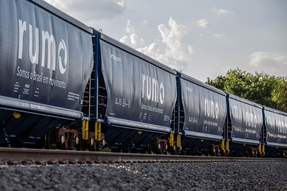 Vagões de trem da Rumo, com a logo e a frase 'Somos o Brasil em movimento' visíveis, alinhados ao longo de uma ferrovia sob um céu parcialmente nublado