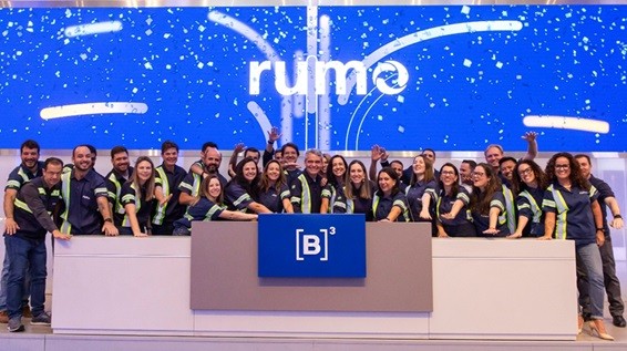 Grupo de pessoas sorridentes em um evento, todas vestindo uniformes em azul e verde. Elas estão reunidas em frente a um painel de fundo iluminado que exibe a palavra 'rumo'. No centro, há um grande painel com o logotipo '[B]³'. Os membros do grupo estão fazendo gestos de celebração, demonstrando entusiasmo e união.