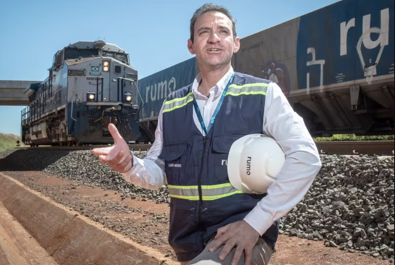 Homem de colete de segurança azul e camisa branca, segurando um capacete branco com a logo "Rumo", posando próximo a uma locomotiva e vagões de trem em uma ferrovia.