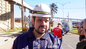 Homem com capacete e uniforme de bombeiro, olhando para a câmera em área externa.