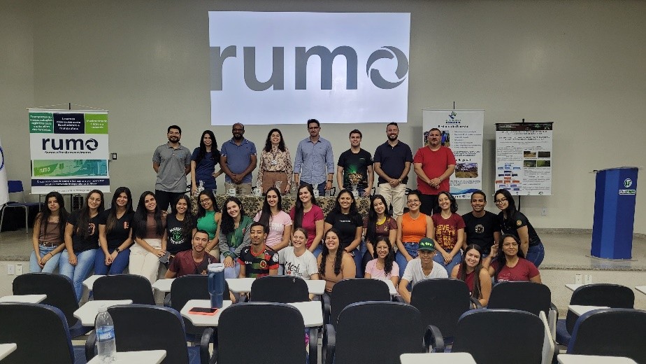 Grupo de pessoas reunidas em um auditório: algumas sentadas nas carteiras à frente e várias em pé sobre o palco, todas posando para a foto diante de uma projeção com o logo "rumo" e banners laterais, em um ambiente de sala de aula/palestra
