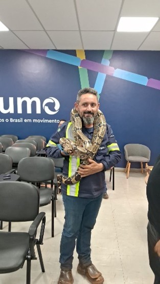 Homem com uniforme de segurança segura uma grande cobra enrolada pelos ombros, em ambiente interno com cadeiras ao fundo.