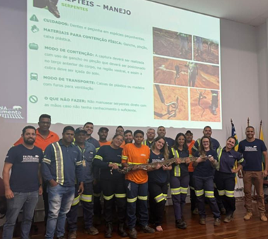 Grupo de participantes e instrutores posando no palco segurando uma cobra comprida durante uma apresentação sobre répteis, com slides projetados ao fundo.