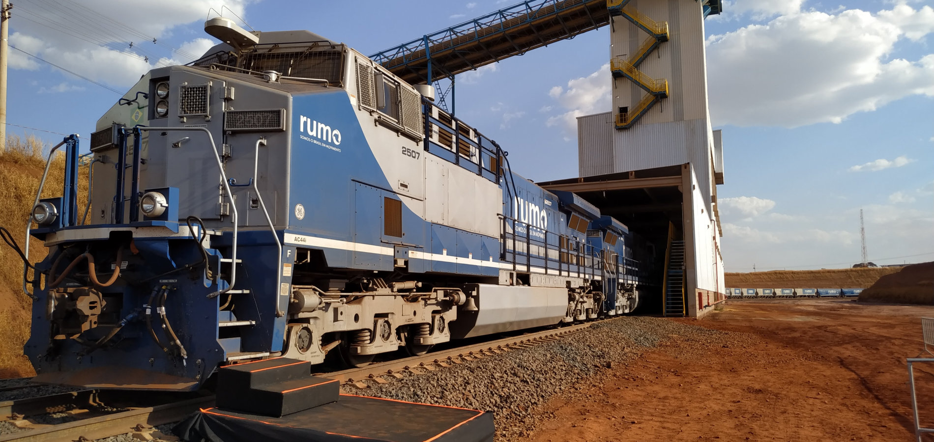 Locomotiva azul da Rumo sobre trilhos diante de uma estrutura industrial de carregamento, sob céu claro.