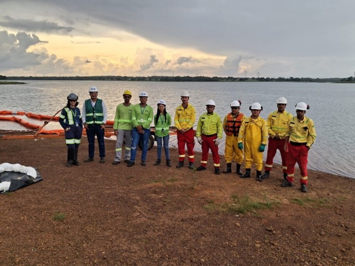 Equipe de segurança com capacetes e roupas de proteção junto à margem de um reservatório, com boias de contenção na água