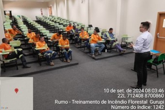 Sala de treinamento com diversos participantes vestindo roupas laranjas sentados em carteiras, com um instrutor em pé na frente.
