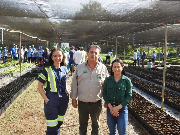 Três pessoas, duas mulheres e um homem, posam para uma foto em uma estufa de mudas, com fileiras de pequenas plantas em sacos pretos ao redor e outras pessoas trabalhando ou observando ao fundo.