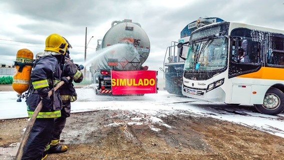 Simulação de acidente de trânsito com dois bombeiros em uniforme e equipamento de proteção operando mangueira de água em frente a caminhão-tanque e ônibus branco e laranja estacionados em piso molhado; placa vermelha com a palavra 'SIMULADO' sobreposta à imagem sob céu nublado.