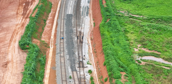 Vista aérea de uma ferrovia em construção, com trilhos parcialmente instalados, equipes e máquinas trabalhando no centro, cercada por áreas de terra vermelha e vegetação verde.