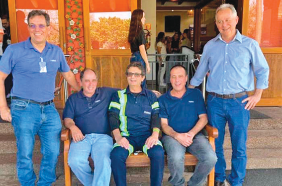 Grupo de cinco homens sorrindo, três sentados em banco de madeira e dois em pé, em ambiente externo
