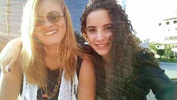 Duas mulheres sorrindo juntas em selfie ao ar livre, com luz solar intensa. Uma tem cabelos loiros lisos e a outra, cabelos cacheados escuros. Ao fundo, estrutura urbana desfocada.