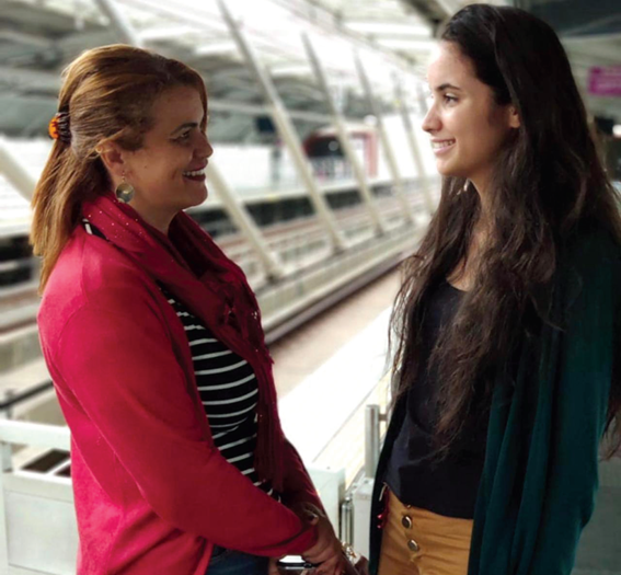 Duas mulheres se olham e sorriem em conversa, em um ambiente amplo com estrutura metálica e vidro ao fundo, que remete a uma estação ferroviária ou terminal. Uma delas usa blazer rosa e a outra, blusa escura.