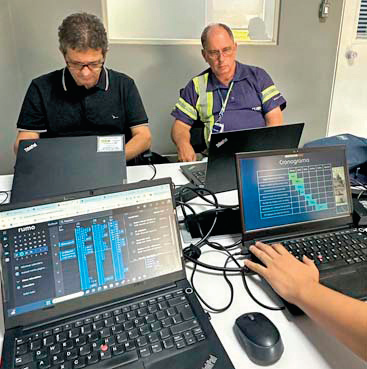 Dois homens sentados em uma mesa de trabalho com três notebooks abertos. Um deles usa colete refletivo e observa a tela. Os monitores exibem sistemas operacionais e um cronograma em apresentação. Ambiente interno de escritório ou sala de operações.