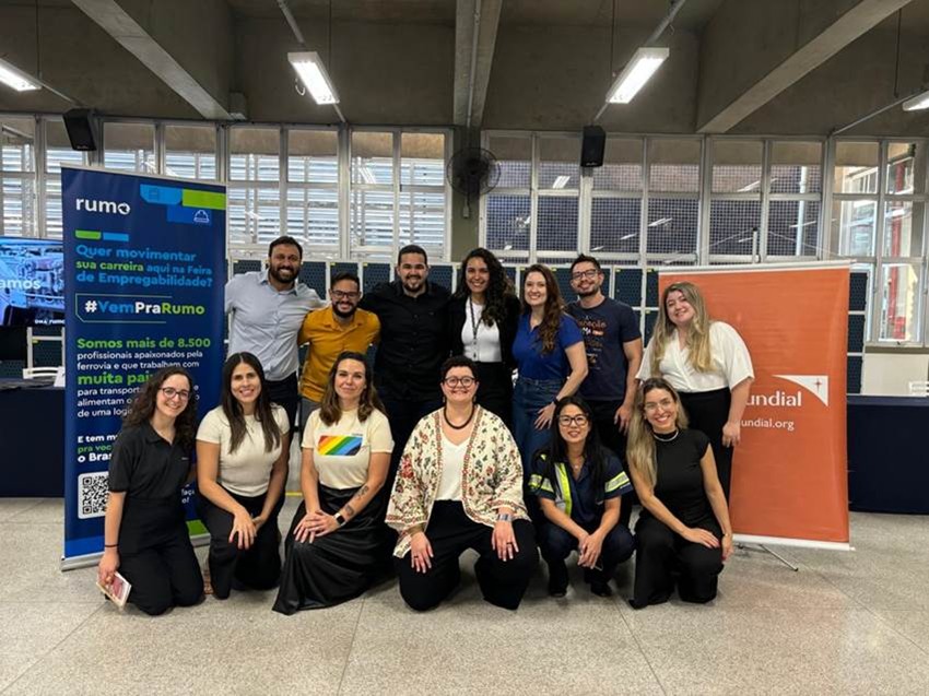 Grupo diversificado de cerca de 13 pessoas sorrindo e posando para foto em um pavilhão ou feira de empregos. Ao lado da equipe, há um banner azul da empresa 'rumo' com a frase 'Quer movimentar sua carreira?' e a hashtag '#VemPraRumo'